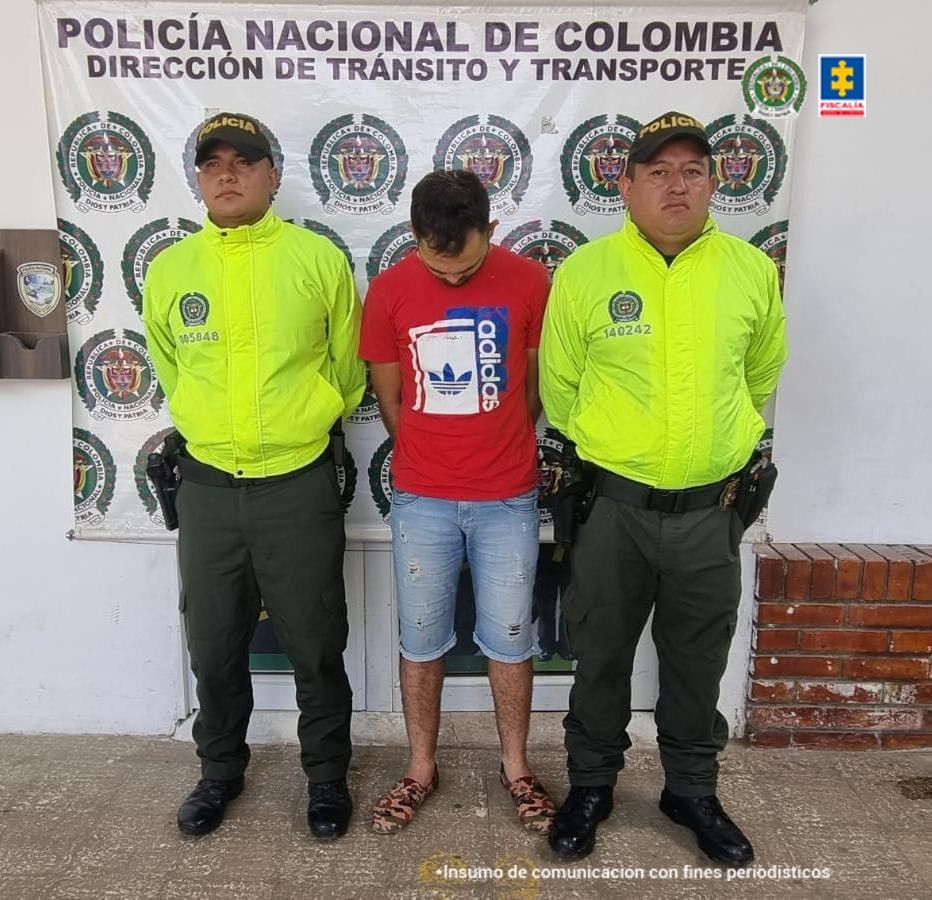 está el capturado de frente manos atrás esposado, custodiado por dos uniformados de la Policía. Detrás banner de la Policía Nacional.