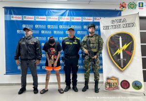 la capturada está de frente esposada y custodiada por un policía, un militar y un investigador del CTI de la Fiscalía. Detrás banner de la Fiscalía y del Gaula Militar.