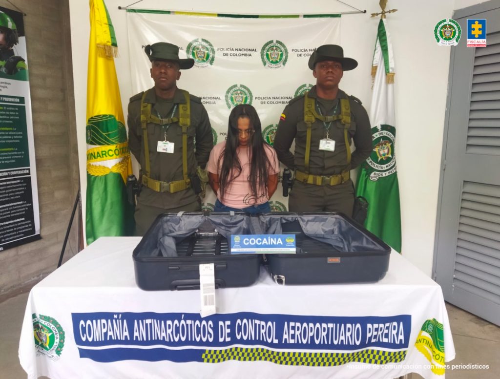 Se observa a una mujer, de frente, y a cada costado un funcionario de Policía Nacional que la custodian. Atrás, un pendón de Policía Nacional. Adelante una mesa en la que se observa la maleta en la cual fue encontrada la sustancia