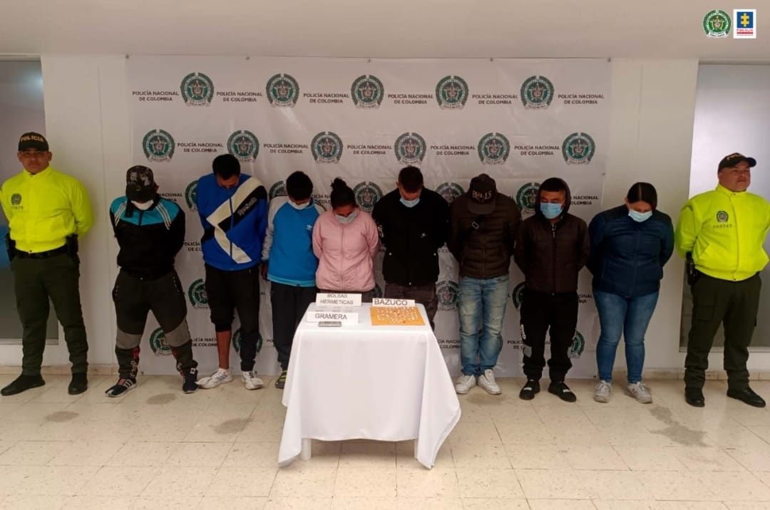En la fotografía se observan a 8 capturados junto a dos agentes de la Policía Nacional. En la parte posterior se observa una mesa con estupefacientes, una gramera y bolsas para empacar drogas