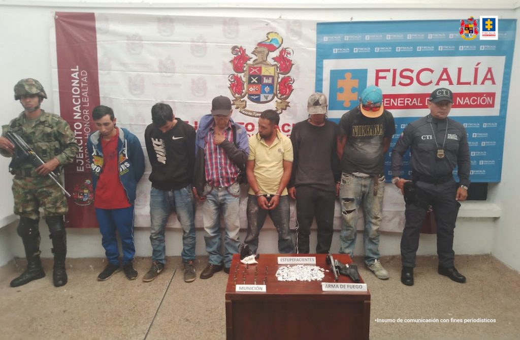 Se observan seis capturados en fila , a un costado un funcionario de CTI y al otro uno de Ejército. Al fondo un pendón de Fiscalía y de Ejército- Al frente una mesa con material incautado en los allanamientos
