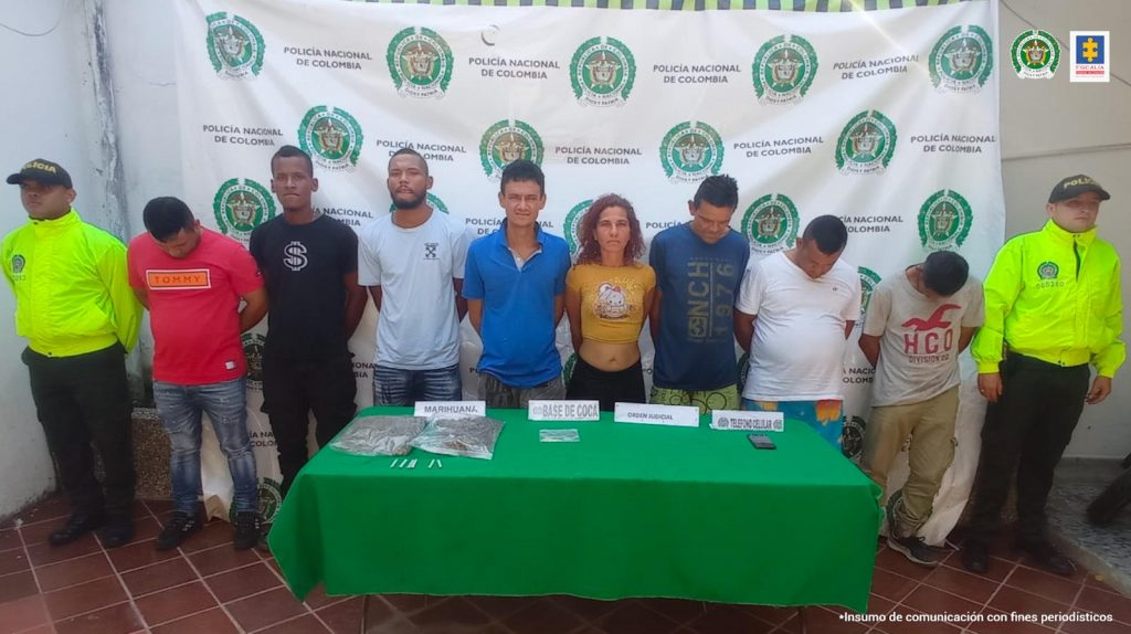 en la foto se aprecia a dos miembros de la Policía Nacional custodiando a los ochos capturados. En una mesa se aprecia los elementos incautados.