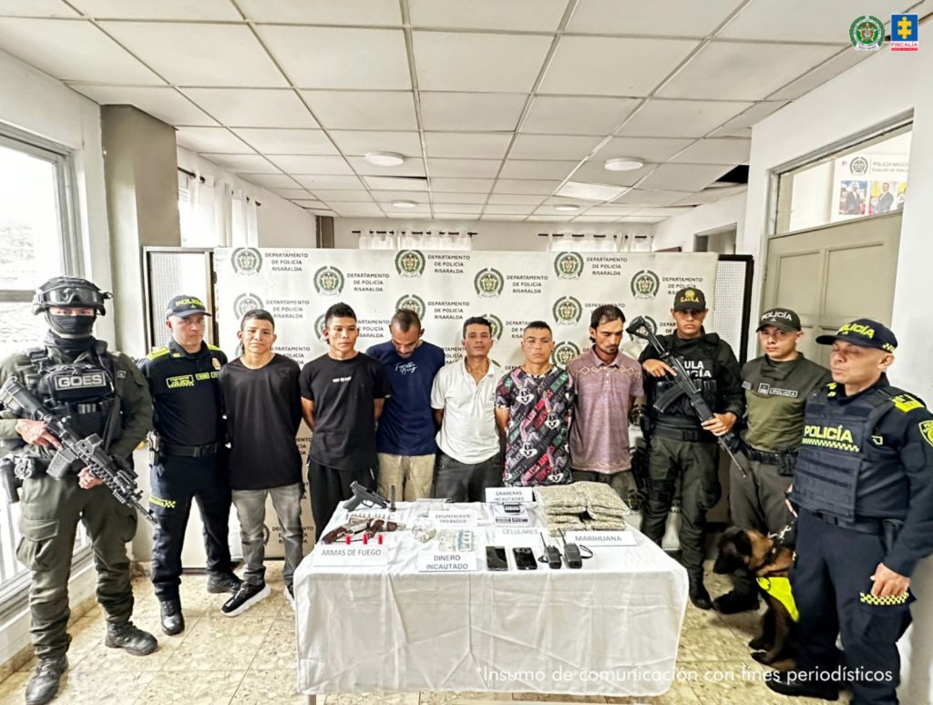 Se observan seis personas capturadas en fila y de frente. A un costado de la fila 3 y al otro dos funcionarios de la Policía Nacional. Adelante , una mesa con el material incautado