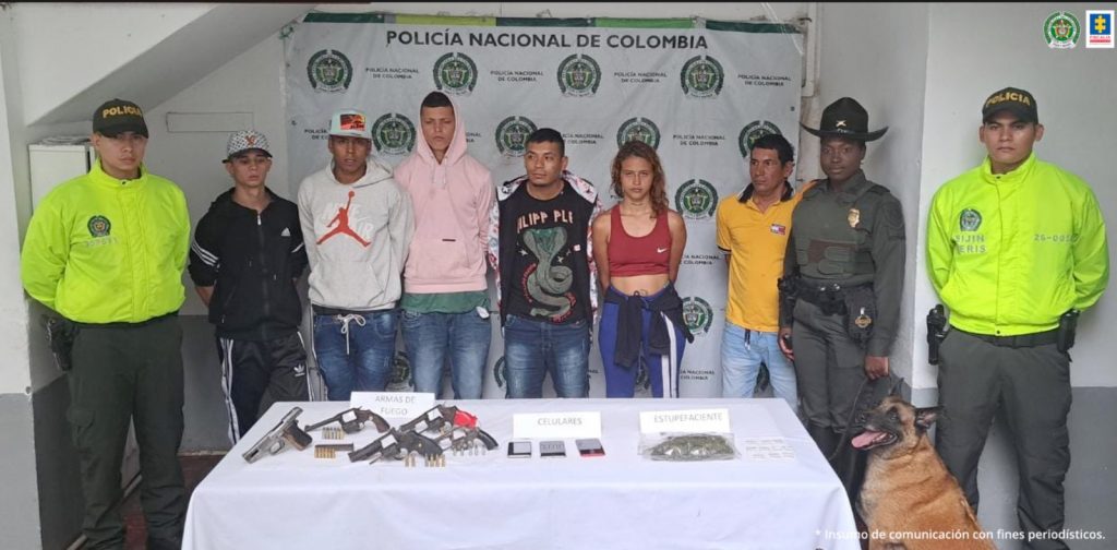 Se observa una fila de seis personas detenidas, a un costado dos funcionarios de policía nacional y un canino de policia nacional. Al otro costado otro funcionario de policía nacional. Al frente una mesa con material incautado. Atrás pendón de Policía Nacional