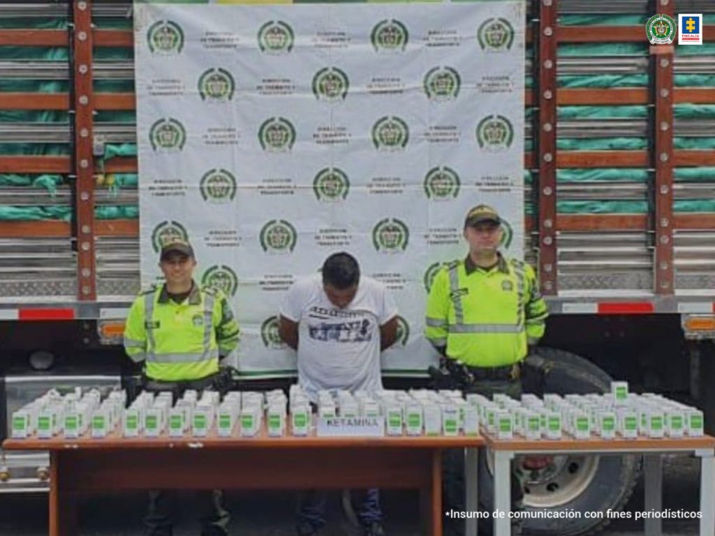 se observa al capturado en el centro, a cada costado un funcionario de Policía Nacional, atrás un pendón de Policía Nacional, sostenido en las barandas del camión en el que, se presume, se transportaba ketamina.  Al frente una mesa con frascos de ketamina que fueron incautados