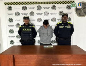 se observa al capturado y a cada costado un funcionario de Policía Nacional, atrás un pendón de Policía Nacional y adelante una mesa con un aviso de que dice "oren judicial"
