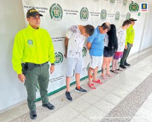 En la fotografía aparecen cuatro capturados junto a personal de Fiscalía Policía Nacional. En la parte posterior de la imagen se ven banners de Policía Nacional y la Fiscalía General de la Nación.
