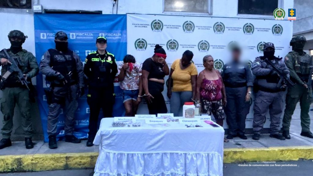 La fotografía fue tomada en exteriores. Se aprecian cuatro mujeres en hilera, esposadas entre sí. A los extremos hay servidores del CTI de la Fiscalía y de la Policía Nacional. Delante de ellas hay una mesa con mantel blanco y sobre ella hay varios letreros que dicen: ‘Marihuana’, ‘Empaques’, ‘Clonazepam’, ‘Celulares’. Asimismo, se aprecian elementos relacionados con dichos avisos. Detrás de las judicializadas hay dos pendones institucionales de la Fiscalía y la Policía.