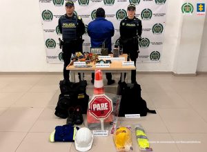 En la fotografía aparece un hombre capturado, acompañado de dos uniformados de la Policía Nacional.  En la parte posterior un banner con logos de la entidad. 