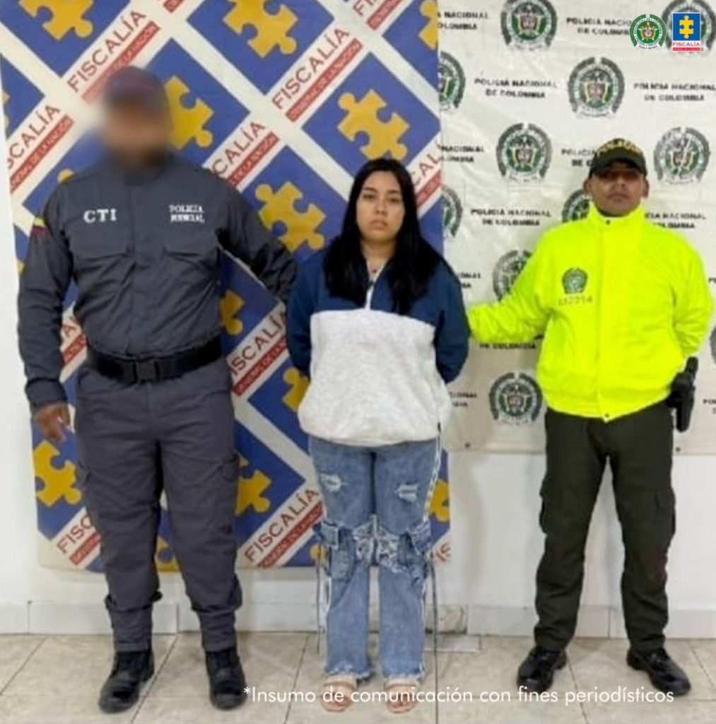 En la fotografía aparece una mujer capturada, acompañada de dos uniformados de la Policía. En la parte posterior un banner con logos de la entidad.