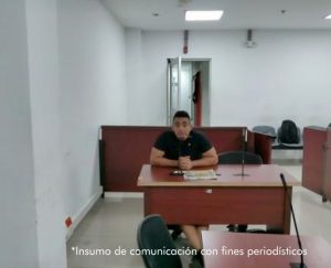 En la fotografía está un hombre de tez trigueña, cabellos negros, vestido con camiseta negrar, sentado en un escritorio de madera con un micrófono en el interior de la sala de audiencia donde se le procesó.