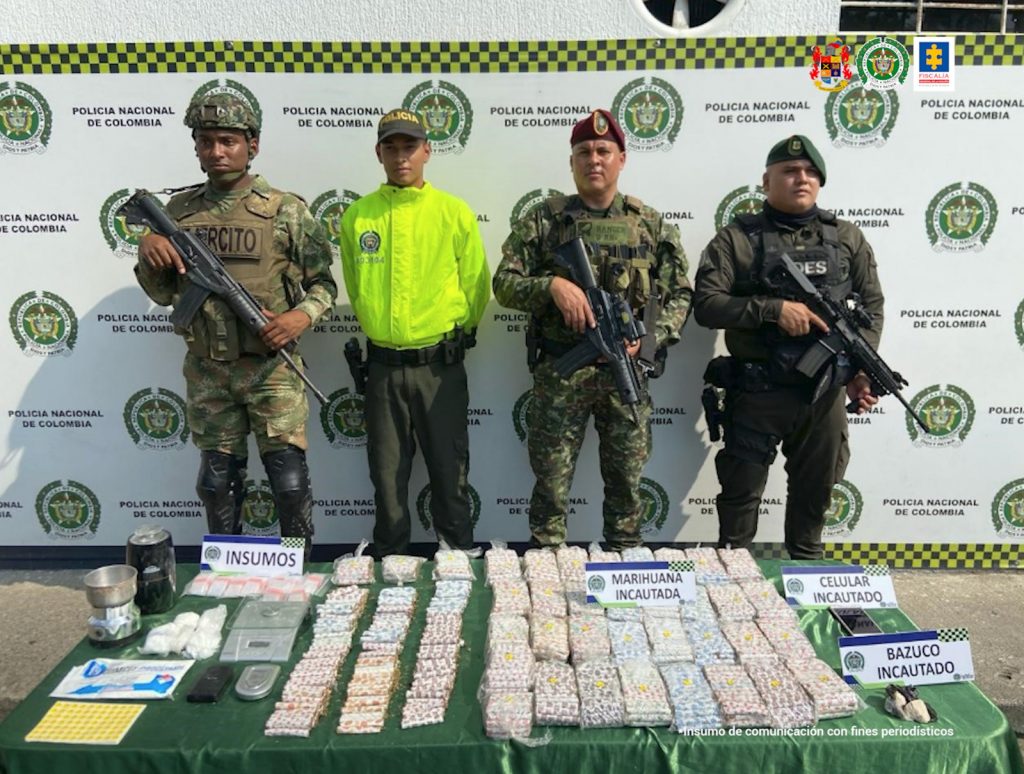 En la fotografía se aprecia a uniformados de la Policía y del Ejército Nacional. Delante de ellos hay una superficie verde y sobre ella hay gran cantidad de bolsas plásticas transparentes, un celular e insumos para el pesaje y embalaje. Asimismo, hay cuatro letreros que dicen: ‘Insumos’, ‘Marihuana incautada’, ‘Celular incautado’, ‘Bazuco incautado’. Detrás de los uniformados hay un pendón institucional de la Policía.