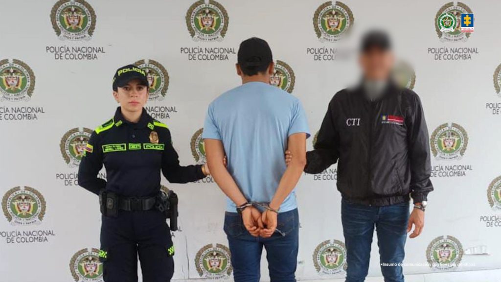 Se visualiza al capturado de espaldas junto a una uniformada de la Policía Nacional y un investigador del CTI de la Fiscalía General de la Nación. Detrás de ellos el banner que identifica a la Policía Nacional.