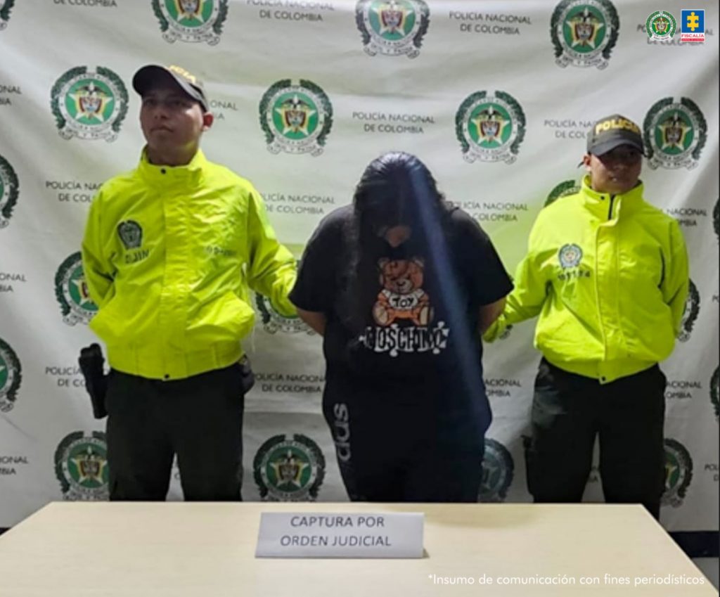 En la fotografía se aprecia de pie, con la cabeza agachada y los brazos atrás a Leidy Johana Osorio Naveros, alias Leidy. Viste con camisa de manga cortal, de color negro y estampadao en el pecho de colores; así como pantalón negro. A sus extremos hay uniformados de la Policía Nacional. Delante de ellos hay una mesa y sobre ella hay un letrero que dice: ‘Capturada por orden judicial’. Detrás de ellos hay un pendón institucional de la Policía.