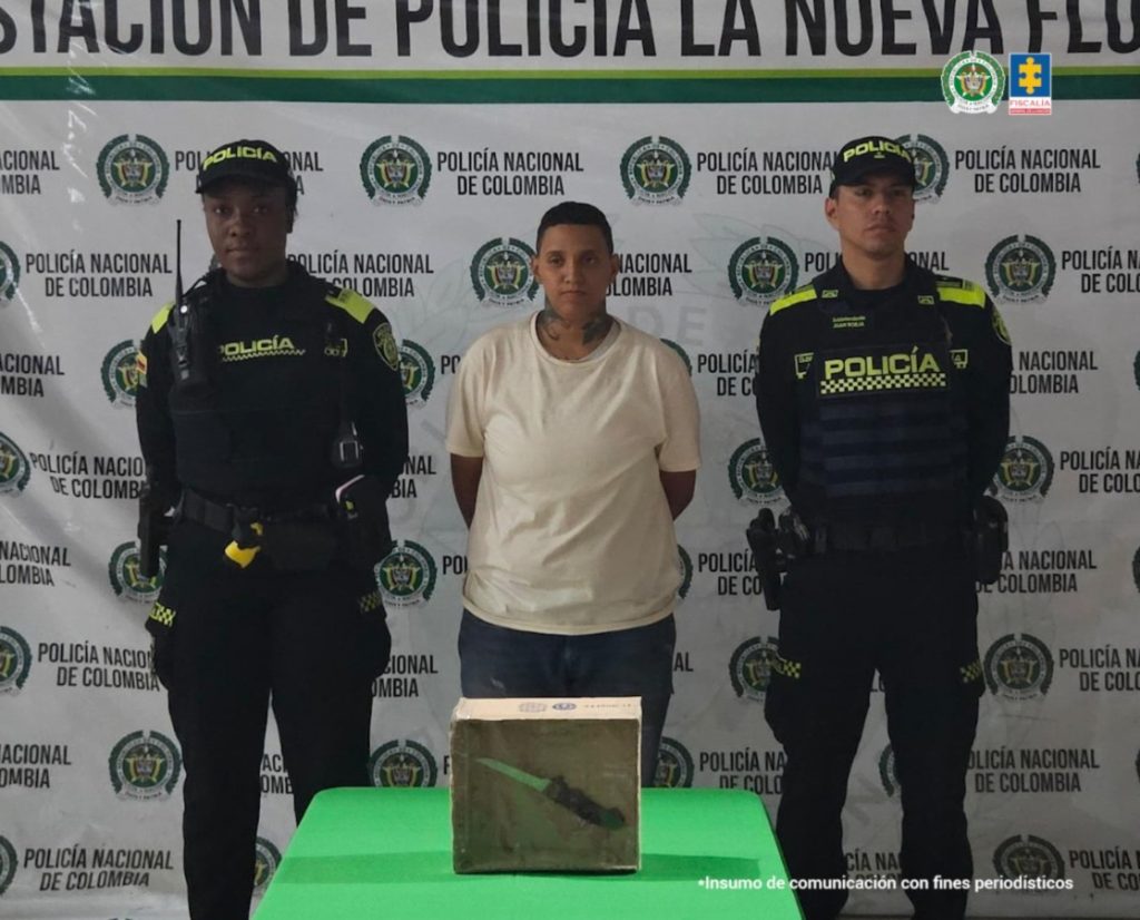 En la fotografía se aprecia de pie y con los brazos atrás a Leidy Tatiana Delgado Mosquera. Usa cabello corto y tatuajes alrededor del cuello. Viste con camiseta de color beage, manga corta y jean azul. A sus extremos hay uniformados de la Policía Nacional. Delante de ellos hay una superficie de color verde y sobre ella hay un contenedor de cartón y se parecia al fondo un cuchillo. Detrás de ellos hay un pendón institucional de la Policía.