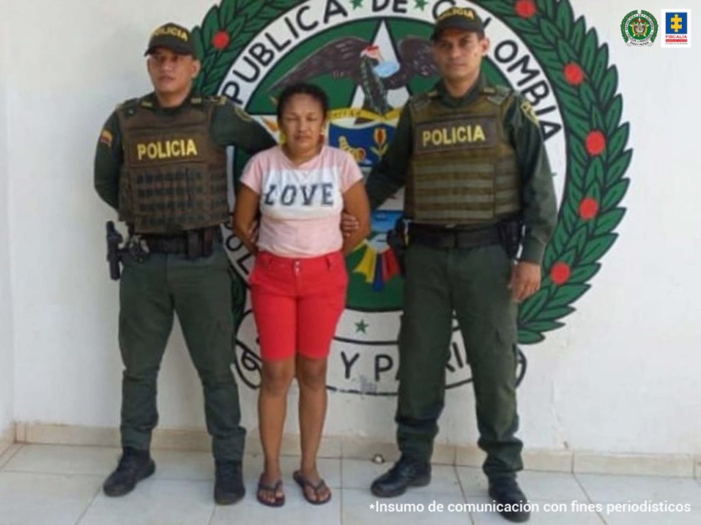 En la fotografía aparece una mujer capturada, junto a dos uniformados de la Policía Nacional. En la parte posterior se encuentra el escudo de la entidad.