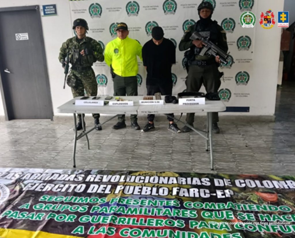 En la fotografía se aprecia de pie, con los brazos atrás y la cabeza agachada a Leonardo Cuellar Villegas. Visten con camiseta, pantalón y zapatillas negras. A sus extremos hay uniformados del Ejército y la Policía Nacional. Delante de ellos hay una mesa y sobre ella hay munición, explosivos, celulares y un porta proveedores. En el piso hay una pancarta de las Farc-Ep. Detrás de ellos hay un pendón institucional de la Policía.
