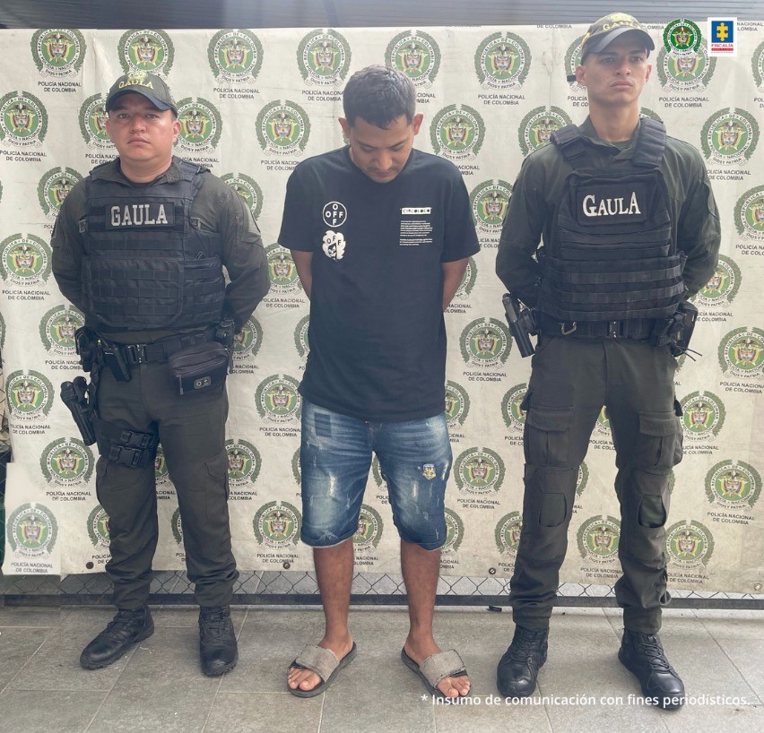 capturado de frente custodiado por dos policías. Detrás banner de la Policía Nacional.