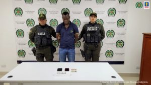 En la foto aparece Leovigildo Mosquera Palacios, alias El Loco o El Compa, con dos funcionarios del Gaula de la Policía Nacional a los lados. Atrás un backing con los logos y adelante una mesa con dos celulares y dos documentos de identificación a los lados.
