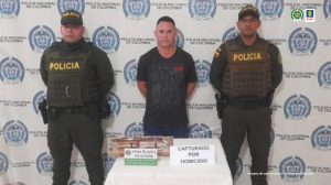 con texto alternativo: En la fotografía aparece un hombre capturado, acompañado de dos uniformados de la Policía. En la parte posterior un banner con logos de la entidad.