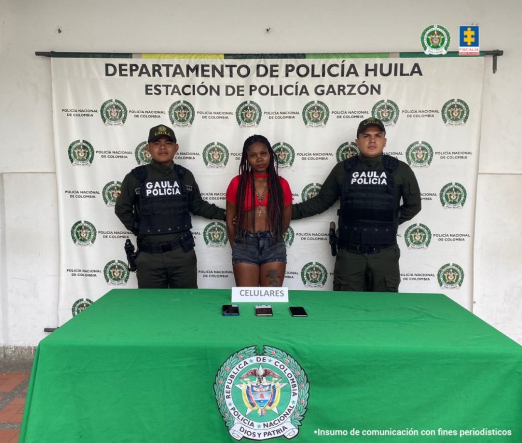 La procesada se encuentra custodiada por dos uniformados de la Sijín de la Policía Nacional. Detrás se observa un backing de la Policía.