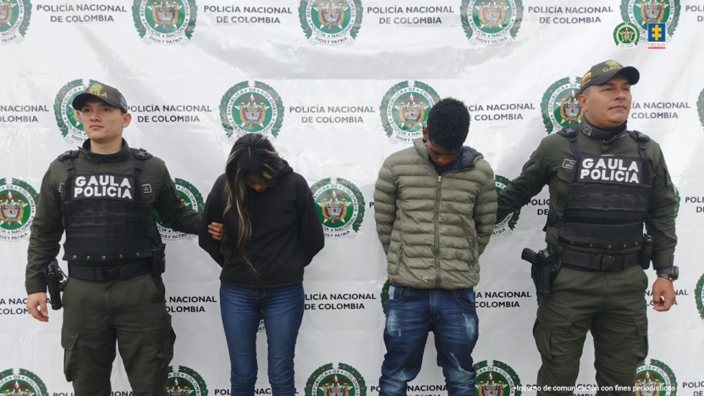 Los capturados se encuentran de frente y con la cabeza agachada y custodiados por dos uniformados del Gaula la Policía Nacional. Detrás de la foto se observa un backing de la Policía Nacional.
