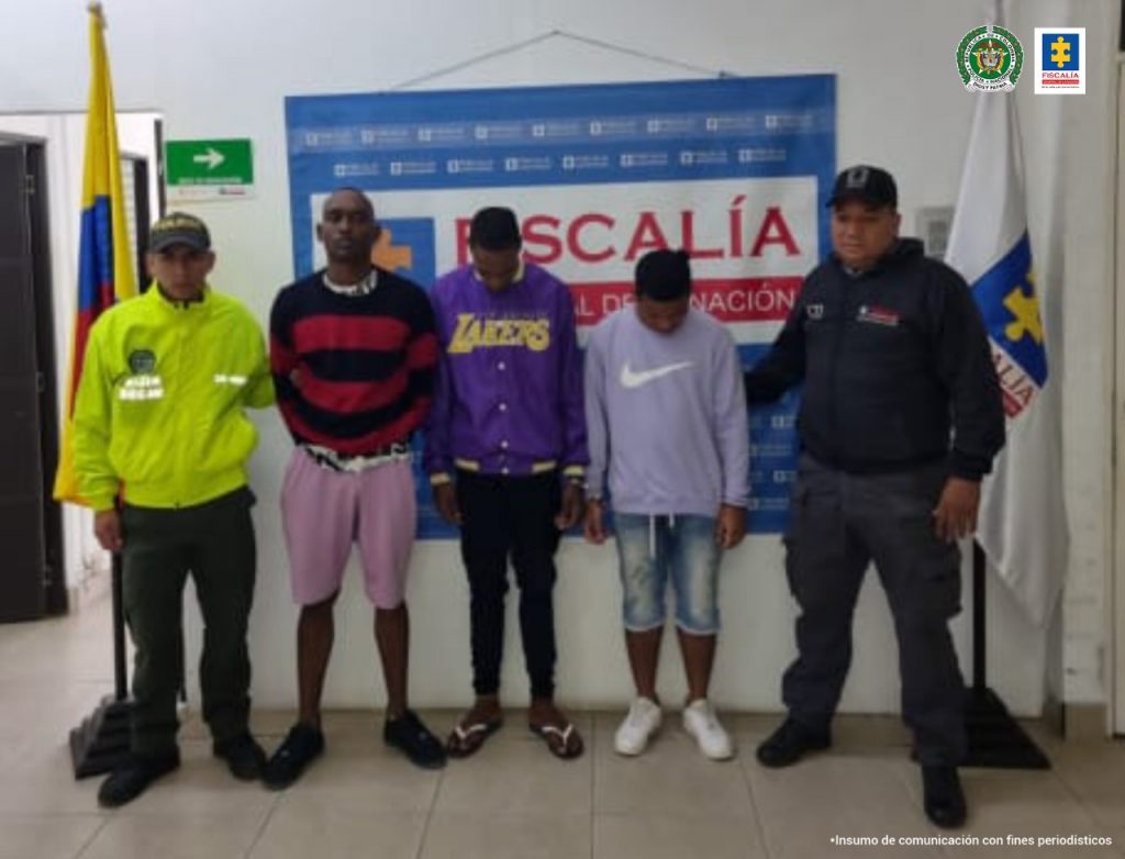 En la foto aparecen de pie los tres presuntos integrantes de ‘Los Aliados’. Al lado derecho de ellos hay un uniformado de la Policía Nacional, junto a la bandera de Colombia. A su izquierda un servidor del CTI de la Fiscalía, junto a la bandera de la Fiscalía. Detrás de ellos hay un pendón institucional de la Fiscalía.
