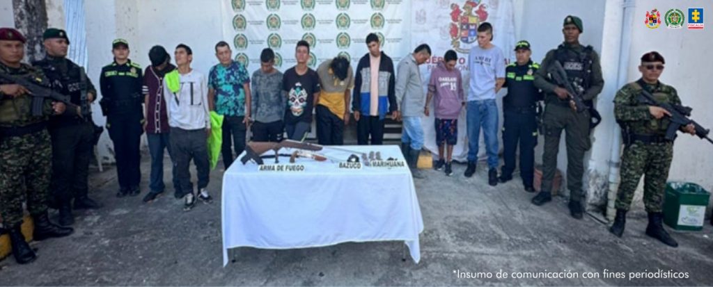 En la fotografía aparecen de pie, en hilera y esposados entre sí los presuntos integrantes del grupo delincuencial Los Centinelas. A sus extremos hay uniformados de la Policía Nacional y del Ejército. Delante de ellos hay una mesa con mantel blanco y sobre ella hay un arma de fuego y bolsas plásticas transparentes. Asimismo, hay tres letreros que dicen: ‘Arma de fuego’, ‘Bazuco’, y ‘Marihuana’. En la parte posterior hay dos pendones institucionales de la Policía y el Ejército.