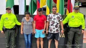  Los tres capturados se encuentran de frente y  custodiados por dos uniformados de la Policía Nacional. Detrás hay unas banderas.