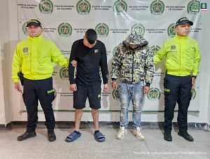 Los imputados se encuentran en custodia por dos uniformados de la Policía Nacional. Uno de los capturados tiene una pantaloneta y chaqueta negra, mientras que el otro tiene un jean con una chaqueta con capucha. Detrás se observa un backing de la Policía.