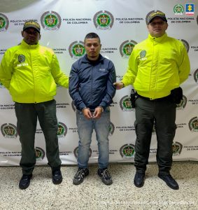 El imputado se encuentra en custodia por dos uniformados de la Policía Nacional, tiene un jean, una chaqueta azul y tennis negros. Detrás se observa un backing de la Policía.