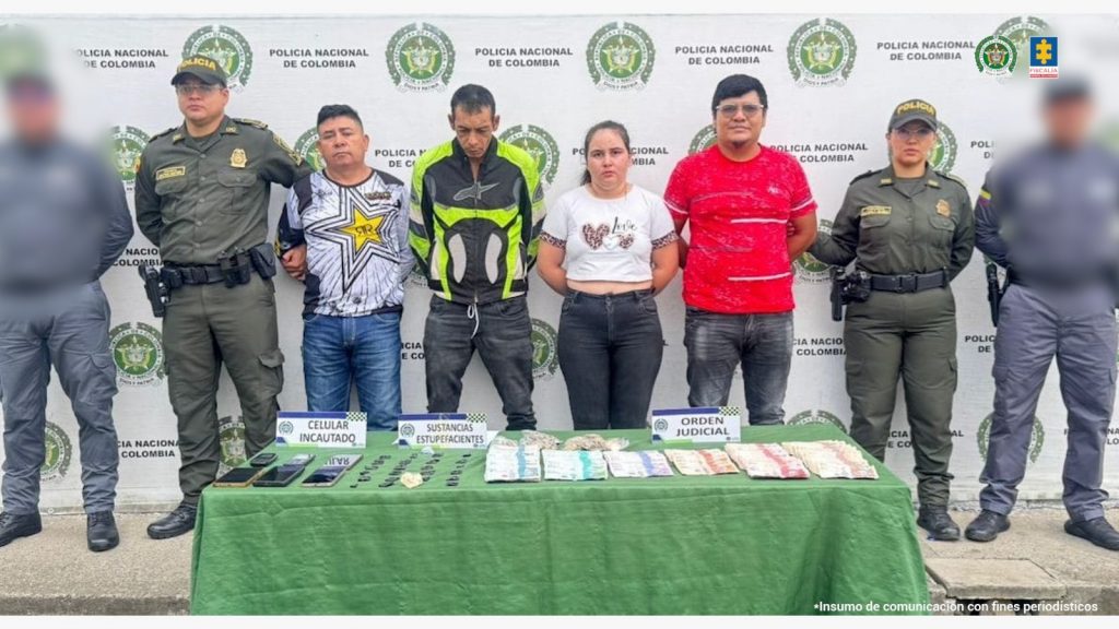 En la fotografía aparecen en hilera y con los brazos atrás los cuatro presuntos integrantes del grupo delincencial Los Express. Hay una mujer y tres hombres. En sus extremos hay servidores del CTI de la Fiscalía y uniformados de la Policía Nacional. Delante de ellos hay una superficie con mantel verde y sobre ella se aprecian celular, bolsas pequeñas transparentes con contenido oscuro y dinero en efectivo. Detrás de ellos hay un pendón institucional de la Policía.
