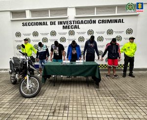 En la fotografía se aprecia en hilera, con la cabeza agachada y esposados a los presuntos integrantes de ‘Los Flets’. A sus extremos hay uniformados de la Policía Nacional. Delante de ellos hay una mesa con mantel verde y sobre ella hay dos letreros, teléfonos celulares y unas bolsas plásticas pequeñas. Asimismo, una motocicleta. Detrás de ellos hay un pendón institucional de la Policía.