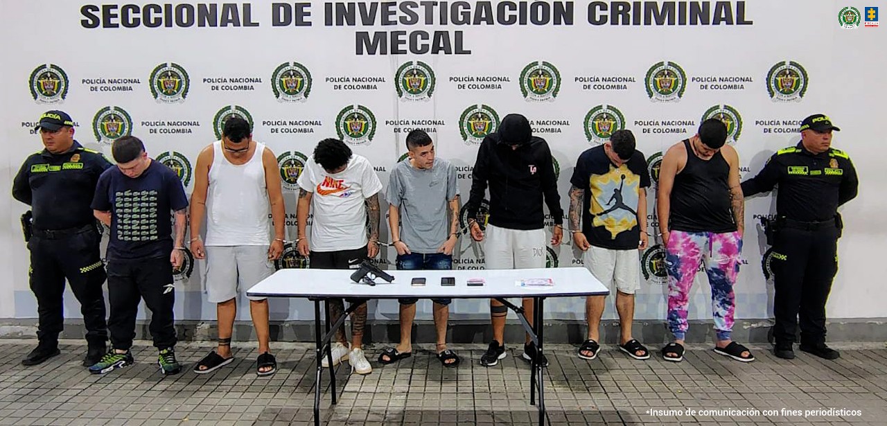En la fotografía se ven de pie, en hilera y esposados entre sí a los presuntos integrantes de ‘Los Jóvenes Millonarios’ o ‘Crypton Fi’. A sus extremos hay uniformados de la Policía Nacional. Delante de ellos hay una pesa rectangular y sobre ellos se aprecian un arma de fuego y celulares. Detrás, hay un pendón institucional de la Policía.