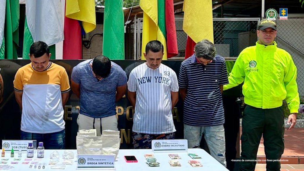 En la foto se observan los cuatro capturados de frente y custodiados por un uniformado de la Policía Nacional. Detrás se observan unas banderas y también se puede apreciar una mesa con parte del material que se incautó durante los allanamientos.