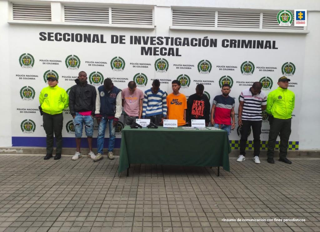 En la foto aparecen de pie los presuntos integrantes el grupo delincuencial Los Panda. A lado y lado de él hay uniformados de la Policía Nacional. Delante de ellos hay una mesa con mantel verde y sobre ell hay dos armas de fuego, munición y unas papeletas. Asimismo, hay tres letreros que dicen: ‘Arma de fuego’, ‘Munición’ y ‘Marihuana’. Detrás de ellos hay un pendón institucional de la Policía.