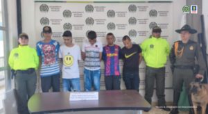 se observan cinco capturados en fila.  En un extremo un funcionario de Policía Nacional y al otro, dos funcionarios de Policía nacional y un canino de Policía Nacional.   Al fondo el pendón de Policía Nacional.
