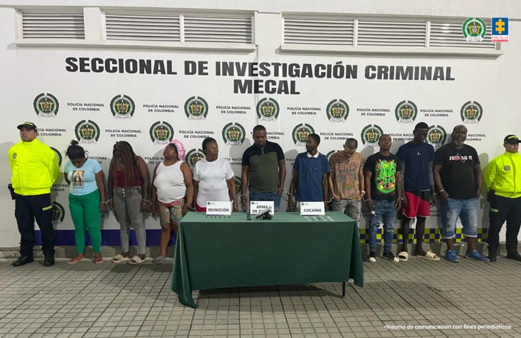 En la fotografía aparecen de pie a los presuntos integrantes del grupo delincuencial Los Raros. A los extremos de ellos hay uniformados de la Policía Nacional. Delante de ellos hay una mesa con mantel verde y sobre ella hay tres letreros que dicen: ‘Munición’, ‘Arma de fuego’, ‘Cocaína’. Asimismo, se aprecia munición, un arma de fuego, un proveedor y varias bolsas plásticas pequeñas con contenido blanco. Detrás de ellos hay un pendón institucional de la Policía.