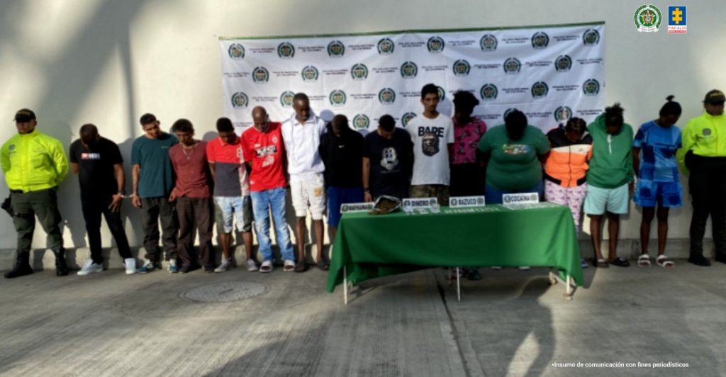 En la fotografía se aprecia en hilera, dentro de un recinto a los 14 presuntos integrantes del grupo delincuencial Los Raros. A los extremos de ellos hay uniformados de la Policía Nacional. Delante de ellos hay una mesa con mantel verde y sobre ella hay cuatro letreros que dicen: ‘Marihuana’, ‘Dinero’, ‘Bazuco’, y ‘Cocaína’. Asimismo, se aprecia una bolsa plástica con contenido verde, dinero en efectivo y bolsas plásticas con contenido blanco.