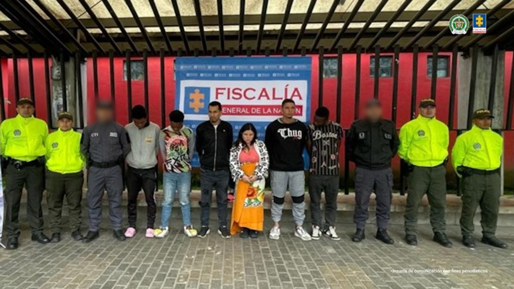 en la imagen se ven los 6 detenidos, 5 hombres y una mujer, al lado de estas personas se encuentran funcionarios del CTI y de la POlicia, atras de ellos hay un baking de la Fiscalia