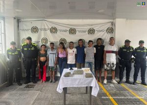 En la imagen se ven varias personas detenidas bajo custodia de integrantes de la Policía Nacional. Delante suyo una mesa con varios elementos incautados. Detrás, un backing institucional.