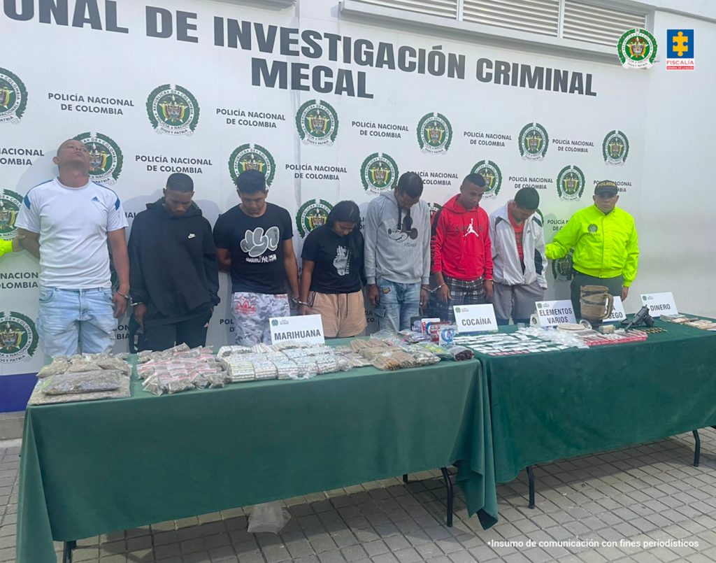 En la fotografía aparecen de pie presuntos integrantes del grupo delincuencial Los del Clan. A los extremos de ellos hay uniformados de la Policía Nacional. Delante de ellos hay dos mesas con mantel verde y sobre ella hay cinco letreros que dicen: ‘Marihuana’, ‘Cocaína’, ‘Elementos varios’, ‘arma de fuego’, ‘Dinero’. Así mismo, se aprecian elementos alusivos a dichos letreros algunos en bolsas plásticas de diferentes tamaños. Detrás de ellos hay un pendón institucional de la Policía.