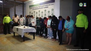 En la fotografía se aprecian de pie, en hilera y con la cabeza agachada a los presuntos integrantes del grupo delincuencial Los del Río. Se encuentran dentro de un recinto. A sus extremos hay uniformados de la Policía Nacional. Delante de ellos hay una mesa con mantel blanco y sobre ella hay dinero en efectivo, un arma de fuego, munición, un celular, bolsas plásticas pequeñas, una bolsa plástica grande con contenido verdoso y unos cigarrillos. Detrás de ellos hay un pendón institucional de la Policía.