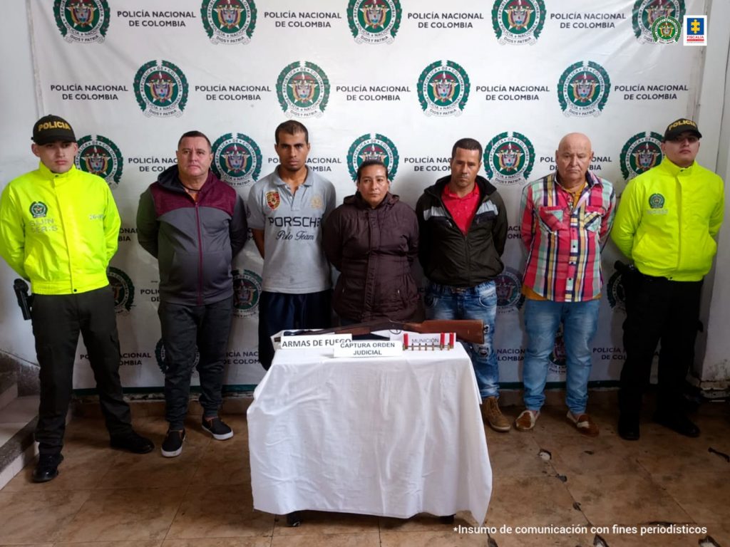 Se observan cinco personas detenidas en fila, cuatro hombres y una mujer, y a cada costado un funcionario de Policía Nacional. Al fondo un pendón de Policía Nacional y al frente una mesa con material incautado.