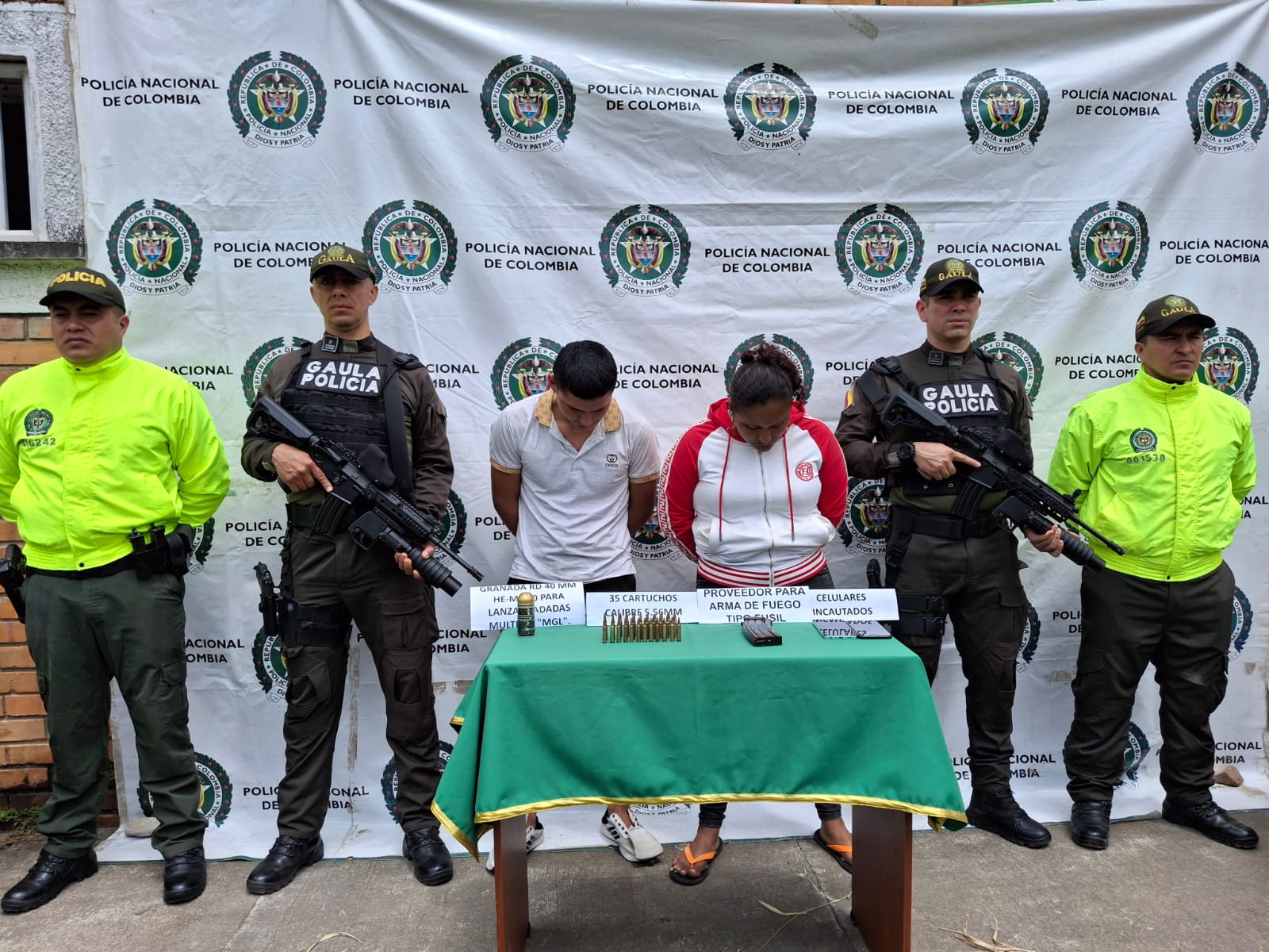 Los capturados se encuentran de frente y custodiados por dos uniformados del Gaula de la Policía Nacional. En la foto se ve de fondo un backing de la Policía, así mismo, se observa una mesa con el material incautado.