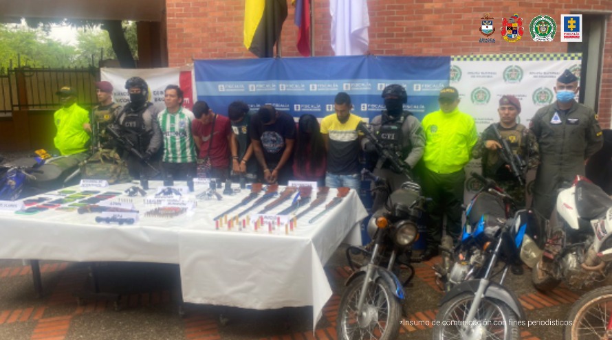 Se visualiza a los seis capturados junto a investigadores del CTI y uniformados de la Policía, Ejército y Fuerza Aérea. Frente a ellos una mesa con las armas incautadas, munición, radios de comunicación, teléfonos celulares y motocicletas. Detrás de ellos los banner institucionales que identifican a la Fiscalía General de la Nación, Policía y Ejército Nacional.