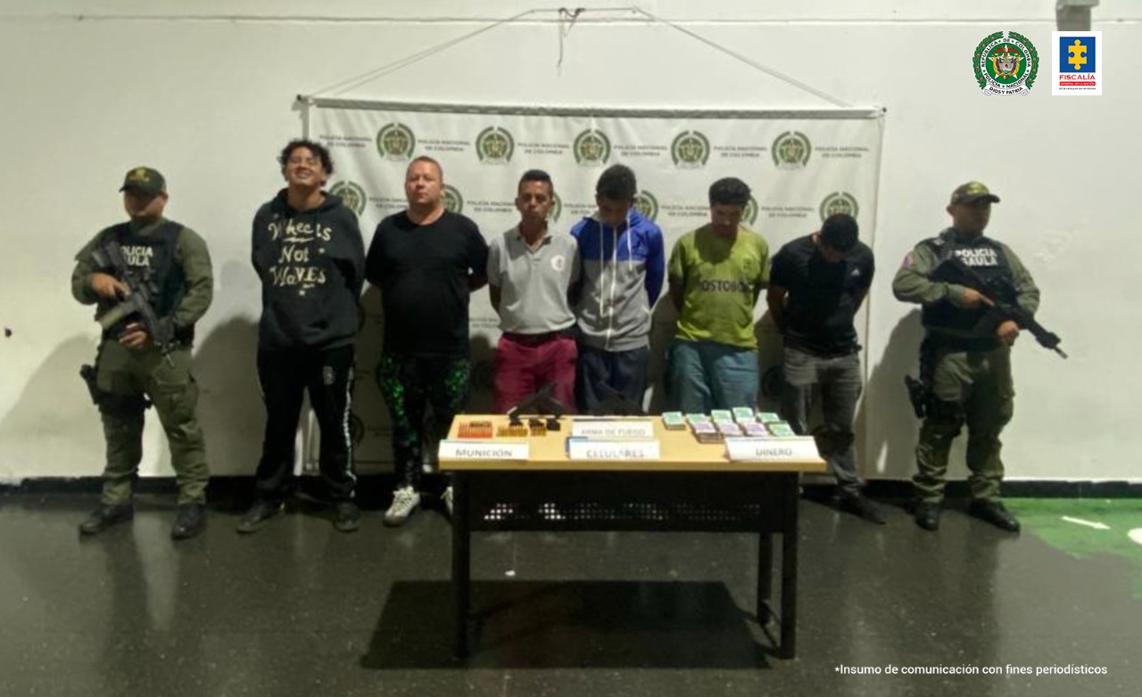 En la imagen se encuentran los 6 procesados con dos servidores del Gaula de la Policía Nacional a los lados y una mesa adelante con los elementos incautados.