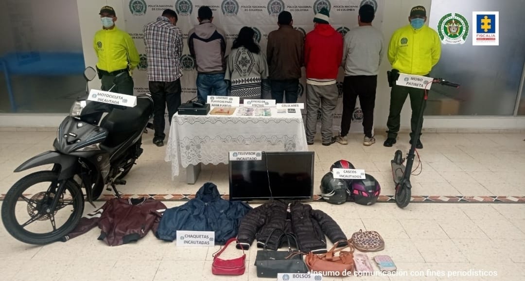 En la fotografía se observan a los 6 capturados junto a dos agentes de la Policía Nacional. En la parte posterior se observa una mesa con dinero, teléfonos celulares, un televisor, chaquetas, bolsos, cascos y una patineta.