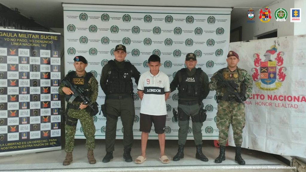 En la imagen se ve una persona de suéter blanco, entre cuatro uniformados de la Policía Nacional, el Ejército y la Armada.