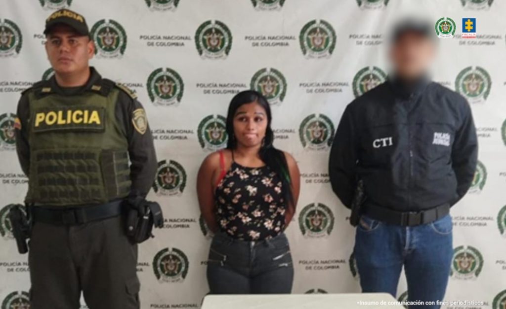En la fotografía se aprecia de pie y con los brazos atrás a Lucy Stella Sánchez Montaño, alias Johana. Viste blusa de color negro con estampado de flores y Jean azul. Usa cabello largo. Al lado izquierdo de ella hay un servidor del CTI de la Fiscalía y a la derecha hay un uniformado de la Policía Nacional. Delante de ellos hay una superficie blanca y sobre ella un letrero que dice: ‘Orden judicial’. Detrás de ellos hay un pendón institucional de la Policía.
